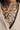 A close-up of a person's neck and chest wearing a gold beaded choker necklace with a heart-shaped pendant, styled with a beige blazer over a black tattooed collarbone.
